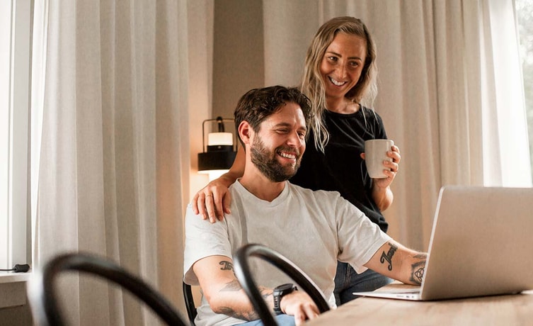 Kvinne holder armen sin rundt en mann som sitter og jobber på PC, til illustrasjon for høyrentekonto