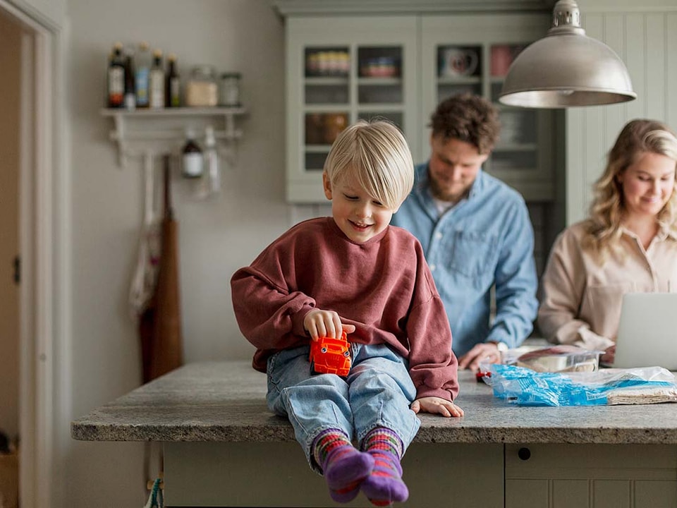 Gutt på kjøkkenbenk, leker med en bil mens foreldrene står bak, til illustrasjon for familieøkonomi