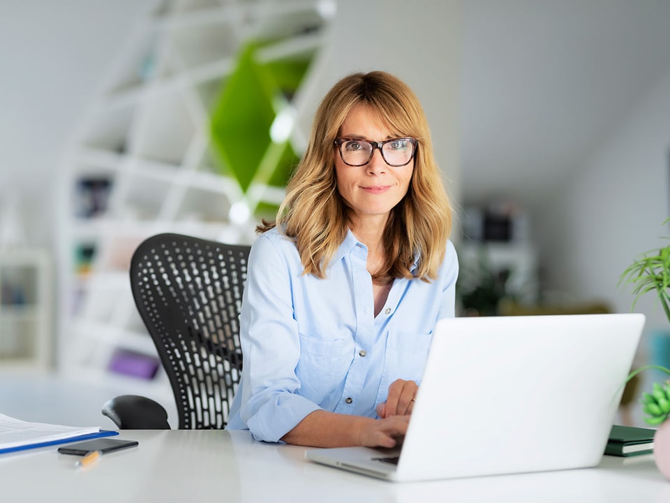 Kvinne med briller og blå skjorte sitter ved en kontorpult og jobber på en laptop, til illustrasjon for betalingstjenester